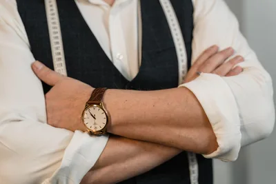 Un sastre cruza los brazos mostrando un elegante reloj de pulsera y una cinta métrica, representando la precisión en la elaboración.